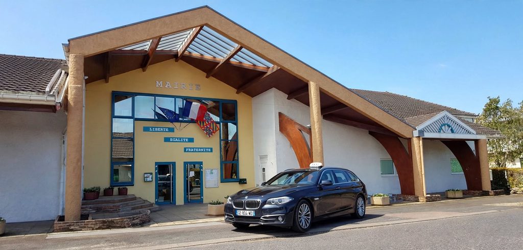 Photographie d'un taxi noir BMW Série 5 Touring break avec enseigne lumineuse TAXI sur le toit, garé devant la mairie d'Oslon en Saône-et-Loire (71). La mairie avec son arche en bois, sa façade jaune marquée MAIRIE, Liberté Égalité Fraternité et ses drapeaux français, européen et bourguignon. Ciel bleu, journée ensoleillée.